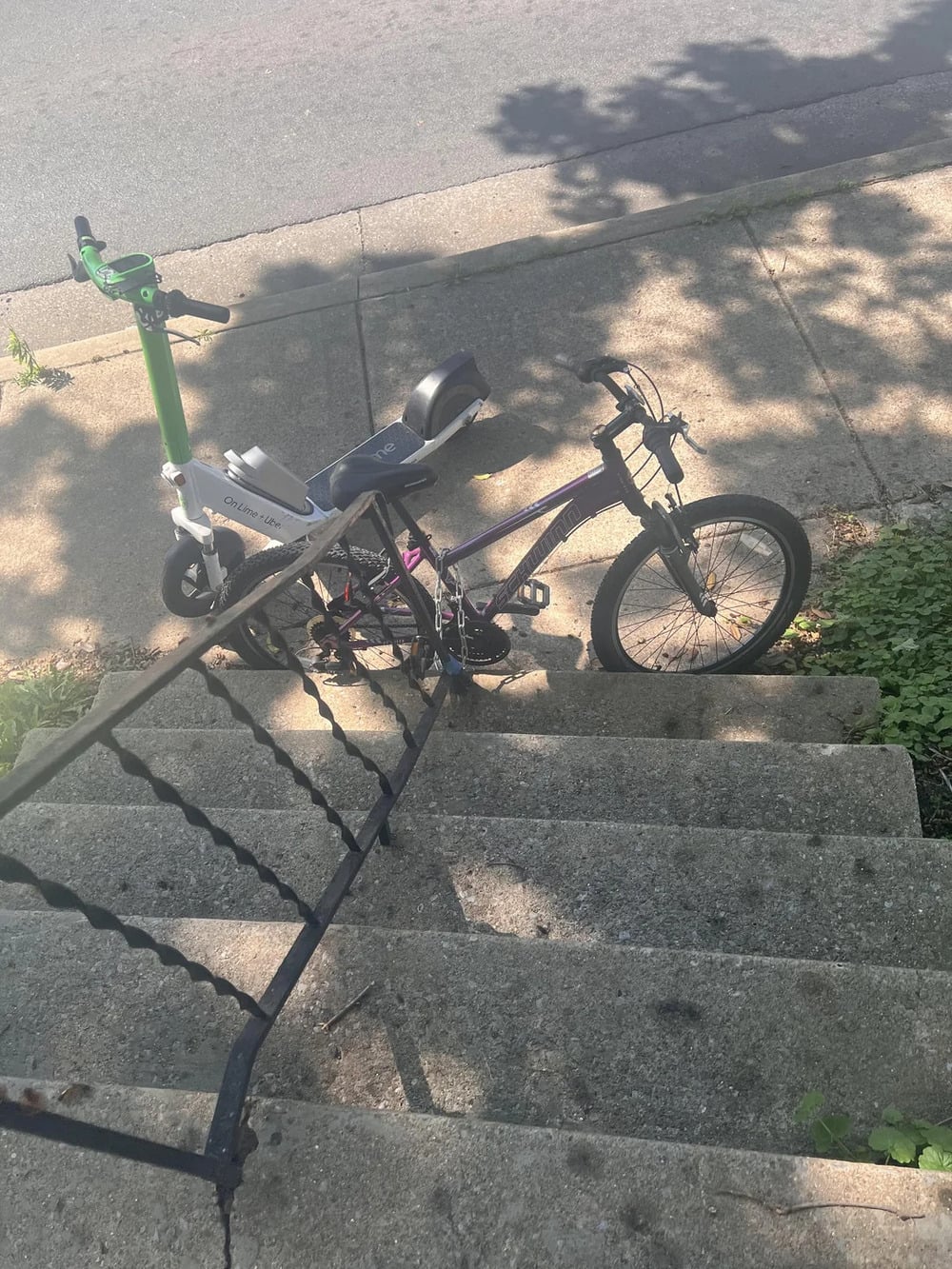 A green electric scooter and a bicycle are locked together at the top of concrete outdoor steps, beside a black metal railing. Sunlight and tree shadows cover the sidewalk and steps.