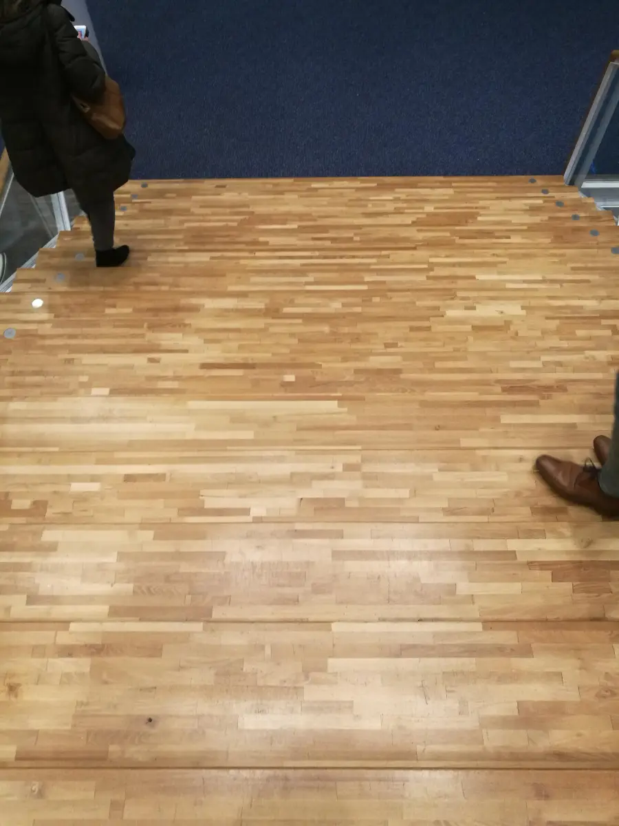 View of a wooden floor with two people at the edges, one wearing black shoes on the left, and the other in brown shoes on the right, both near a blue carpeted area.