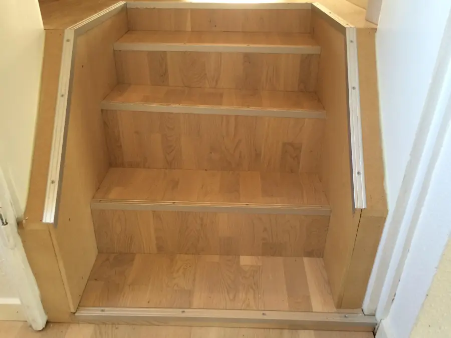 Wooden staircase with four steps, each step becoming progressively deeper from top to bottom. The stairs are bordered by light-colored wooden paneling with metal edge trims on both sides.
