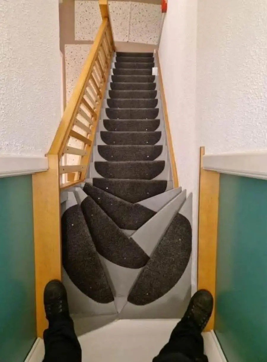 A person’s feet are visible at the top of a staircase with oddly shaped, uneven black and gray steps, making the stairs appear difficult and unsafe to walk down.