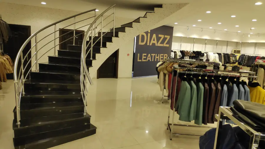 A clothing store with a black staircase, metal railing, racks of jackets, and a wall sign that reads “DIAZZ LEATHER.” The floor is shiny, and the store is brightly lit.
