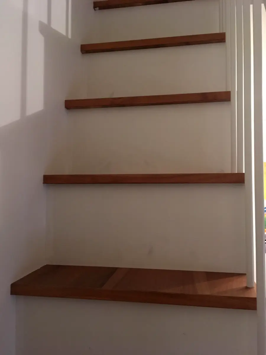 Wooden stairs with no risers seen from below, attached to a white wall with vertical white railing bars on the right side. The lighting creates soft shadows on the wall and the steps.