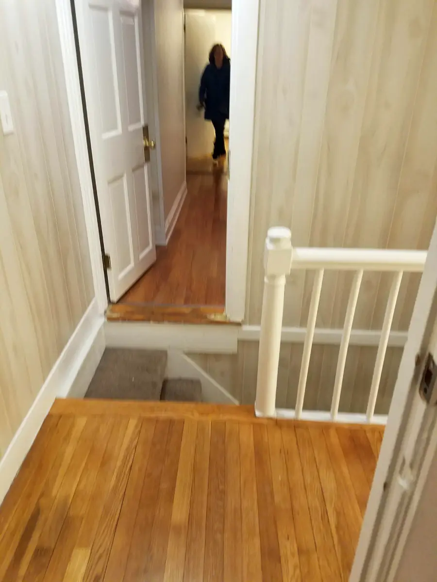 A hallway with wooden floors leads to a doorway with a sudden step down. Stairs with a white railing are visible. A person stands in the background near another door.