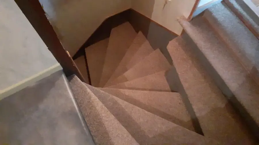 Carpeted spiral staircase taken from above, showing a tight turn and several angled steps leading down to another level. The stairs are bordered by walls and a railing.
