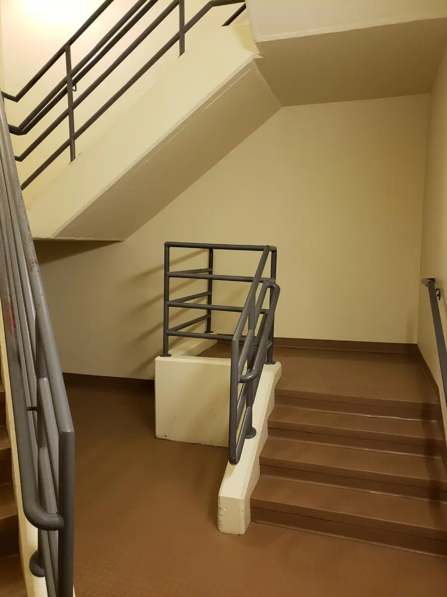 Interior stairwell with brown steps, beige walls, and gray metal railings. The stairs split directions at a small landing beneath an upper flight. The area is well-lit with no visible decorations or windows.