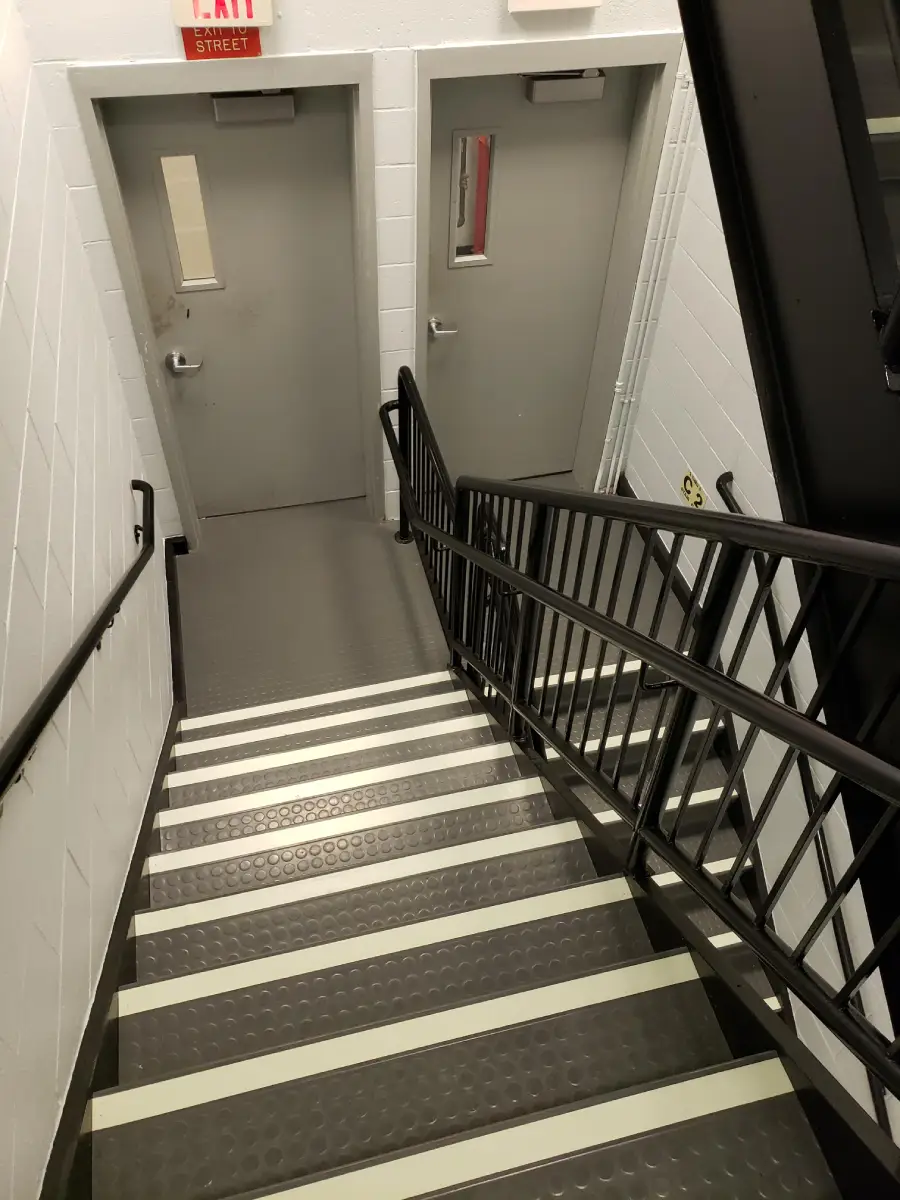 A gray indoor stairwell with black railings, white-striped steps, and two closed gray doors at the bottom. An exit sign is visible above one door. The walls are white and the area is well-lit.