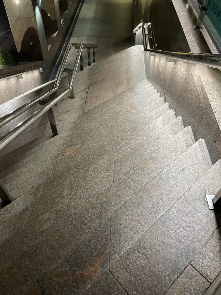 A steep set of marble stairs with metal railings on both sides, leading down to a dimly lit underground area. The lighting creates reflections on the smooth stone surface.