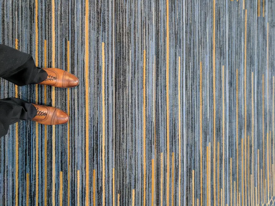 A person wearing brown dress shoes and black pants stands on a blue, gray, and yellow striped carpet.