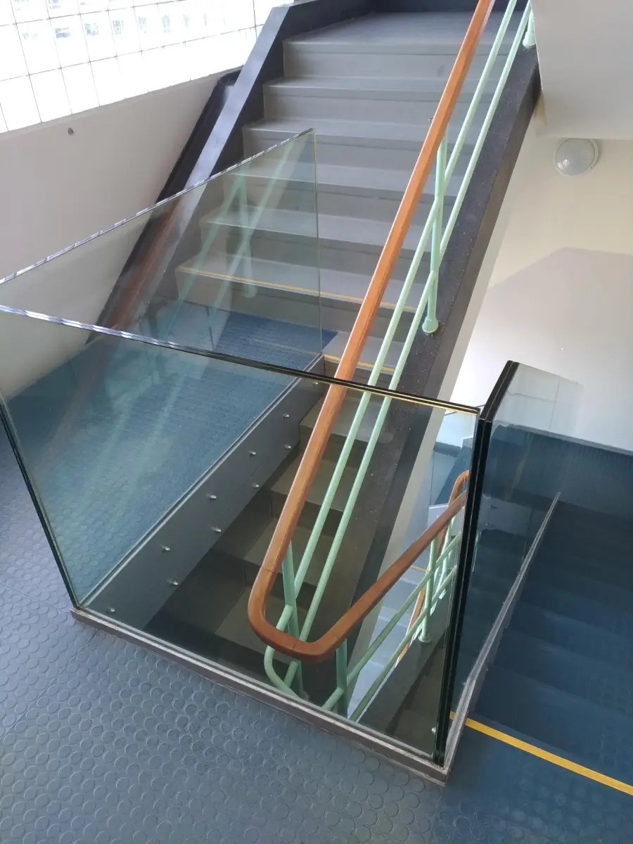 A staircase with glass panels on the sides and a wooden handrail, viewed from above. The stairs have gray steps and light blue railings, and the floor is covered with blue hexagonal tiles.