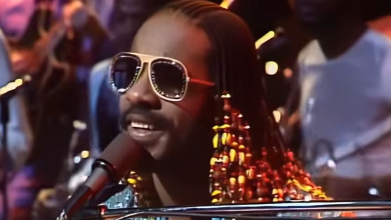 A musician with long, beaded hair and sunglasses sings into a microphone while playing the keyboard on stage, with band members blurred in the background.
