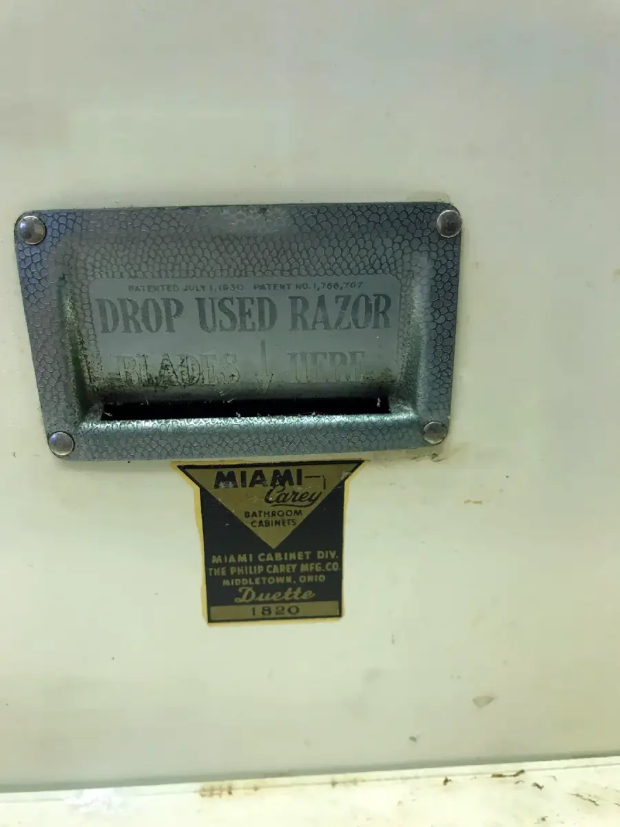 Metal slot labeled “Drop Used Razor Blades Here” on a bathroom cabinet. Below is a yellow and black sticker reading “Miami Carey Bathroom Cabinet” with additional manufacturer information.