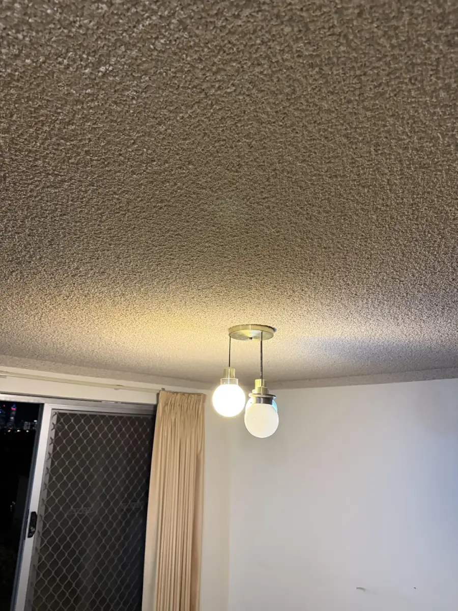 Two light bulbs hang from a ceiling fixture; one bulb is lit and the other is off. The ceiling has a textured finish. A sliding glass door with a curtain is visible in the background.