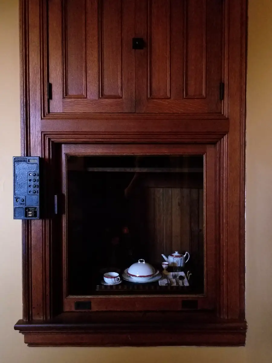 A wooden dumbwaiter with a glass window holds a tray with a teapot, a cup and saucer, a covered dish, and a creamer inside. A control panel is mounted on the left side of the frame.