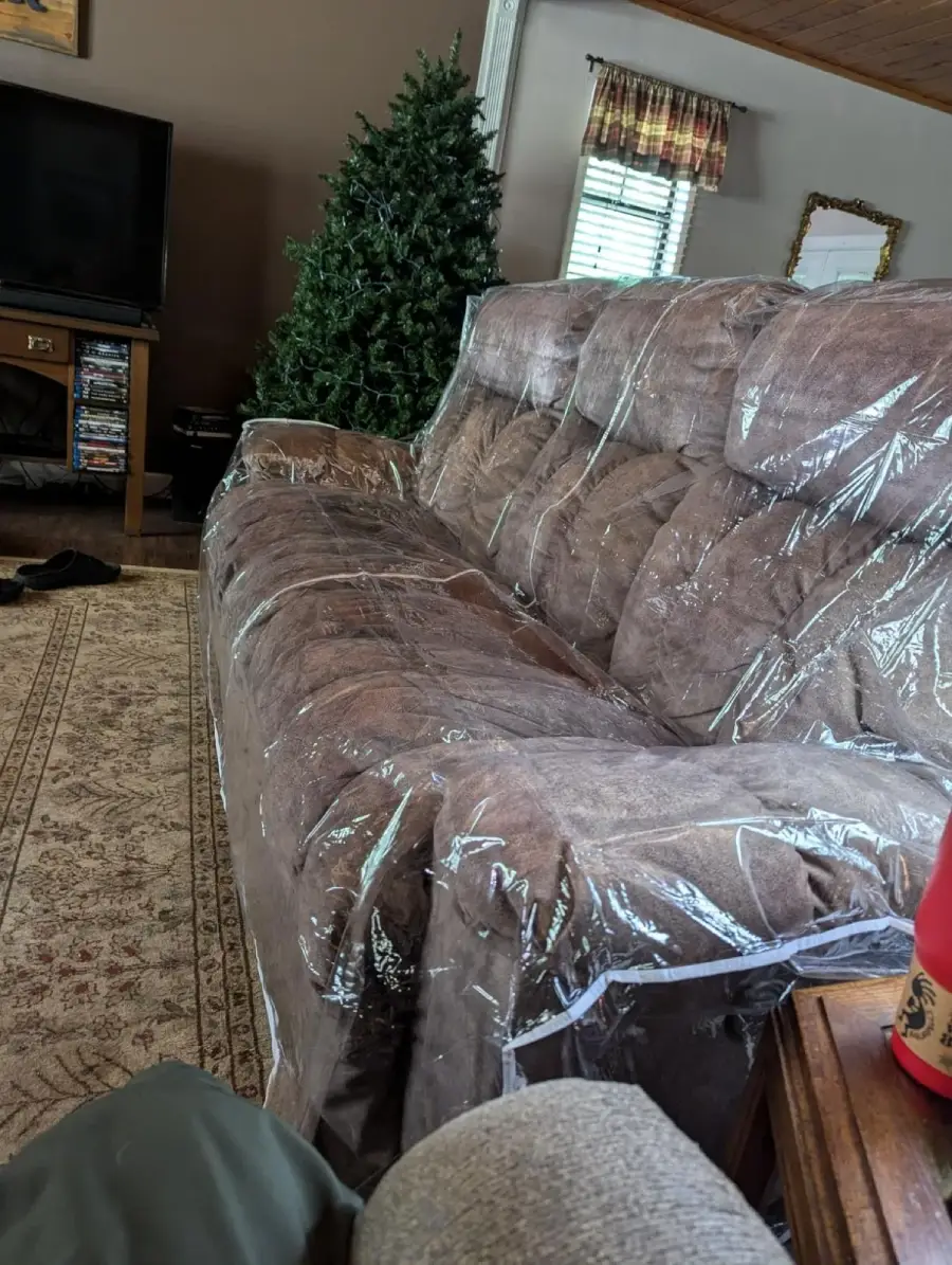 A brown couch covered in clear plastic sits in a living room with a rug, an unlit Christmas tree, a TV, and a side table with a red drink bottle.