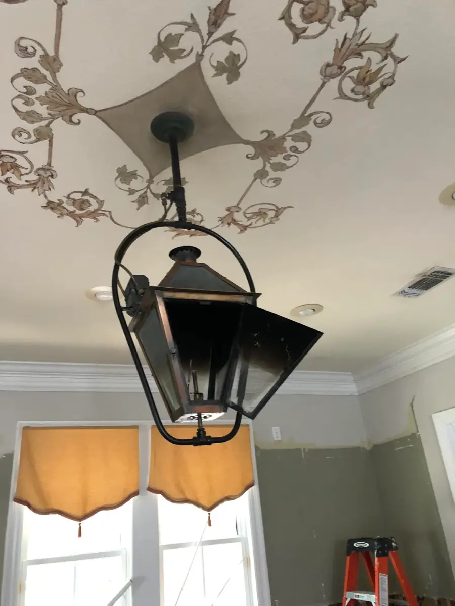 A black lantern-style ceiling light is installed upside down in a room with ornate ceiling decor, yellow window shades, and a red ladder partially visible in the corner.