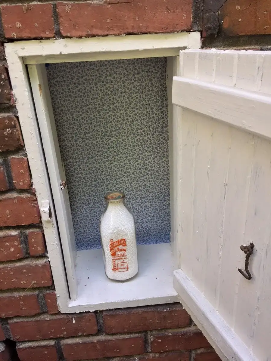 A glass milk bottle sits inside a small, open white cupboard built into a brick wall. The cupboard has a patterned blue and white interior.