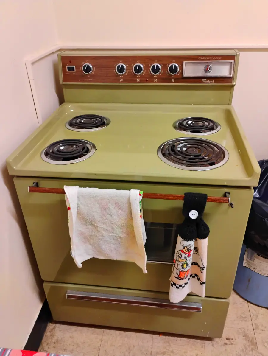 A vintage olive-green electric stove with four coil burners, a wooden control panel with knobs, and two towels hanging from the oven handle in a small kitchen space.