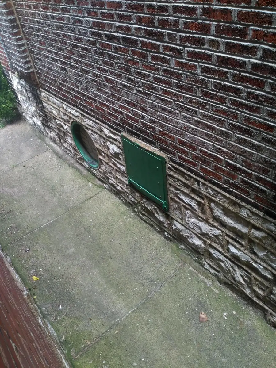 A narrow sidewalk runs alongside a brick and stone wall with a round green-framed window and a rectangular green metal panel.