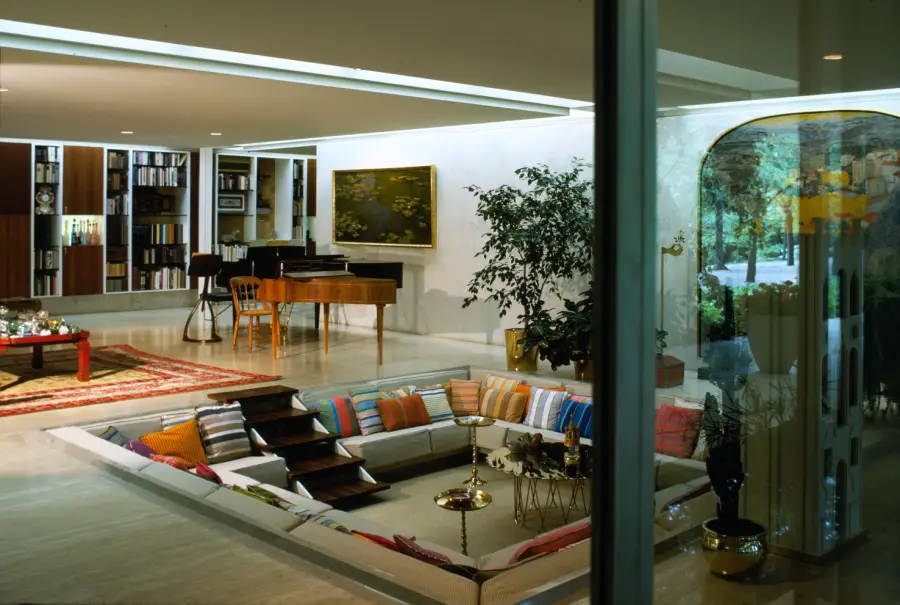 A stylish sunken living room with colorful pillows, a central table, and brass décor, adjacent to a grand piano, bookshelves, and large windows overlooking greenery.