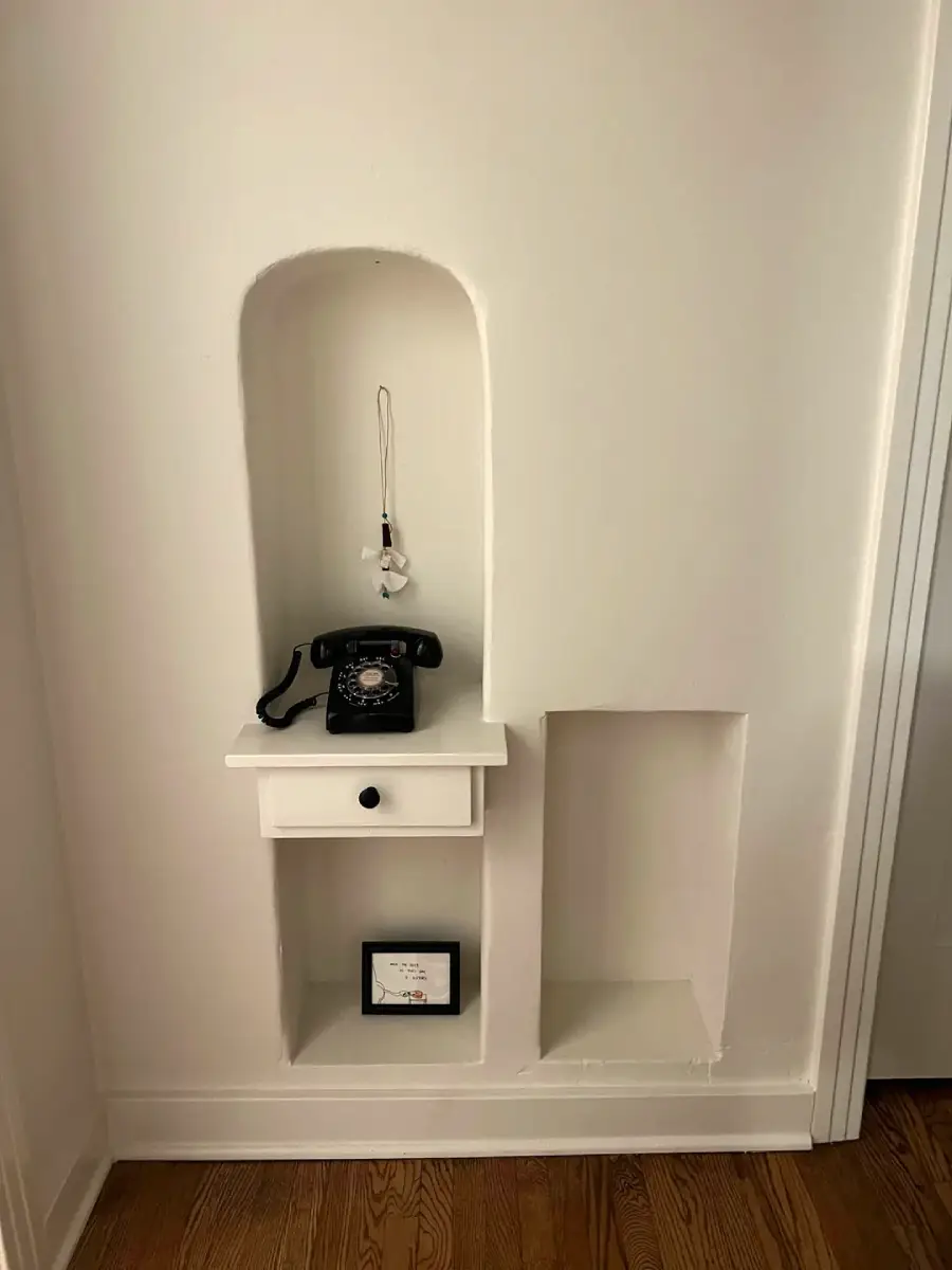A vintage rotary phone sits on a small white shelf with a drawer; above it hangs a keychain. Below the shelf is a recessed niche with a framed photo and another empty, larger niche beside it.