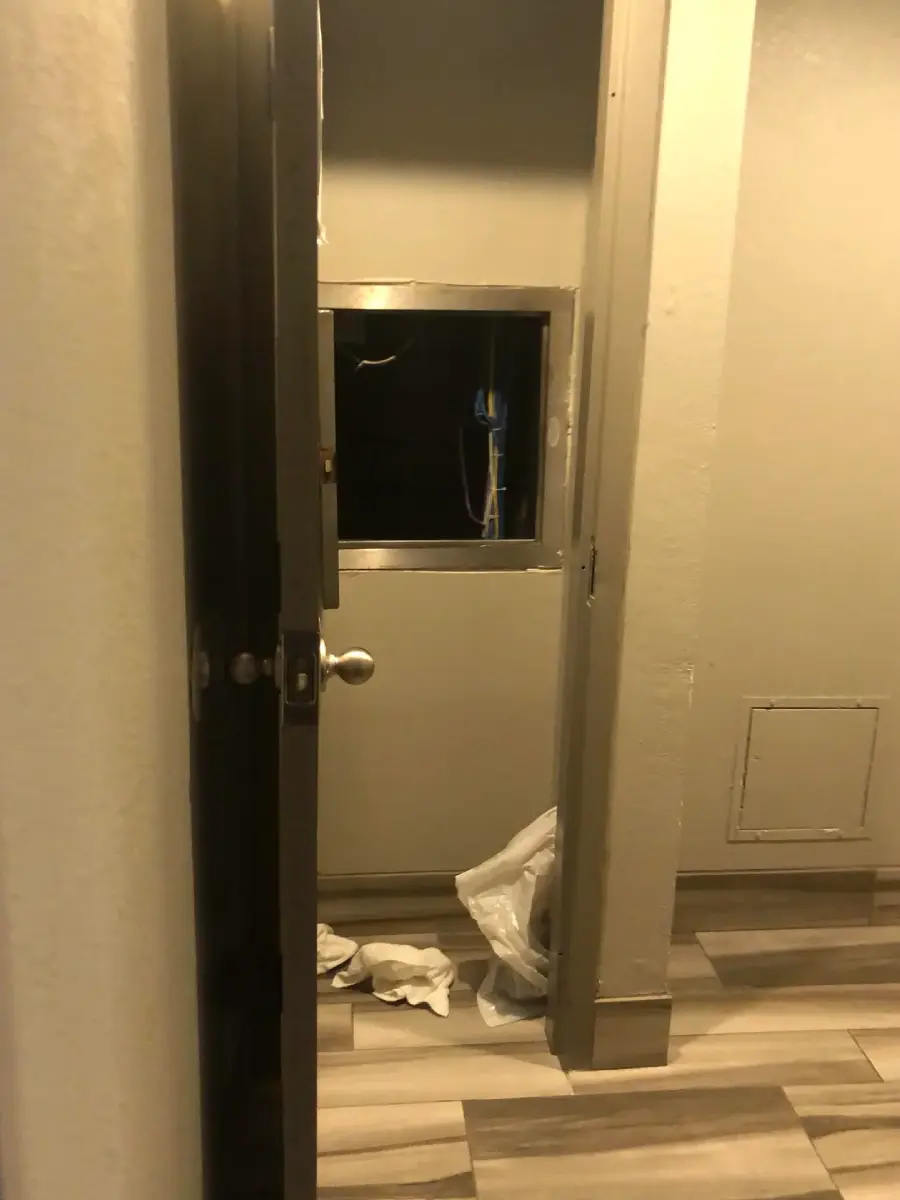 A small room with beige walls, a window with a mop visible through it, tiled floor, and some crumpled tissues or paper towels on the ground near the door.