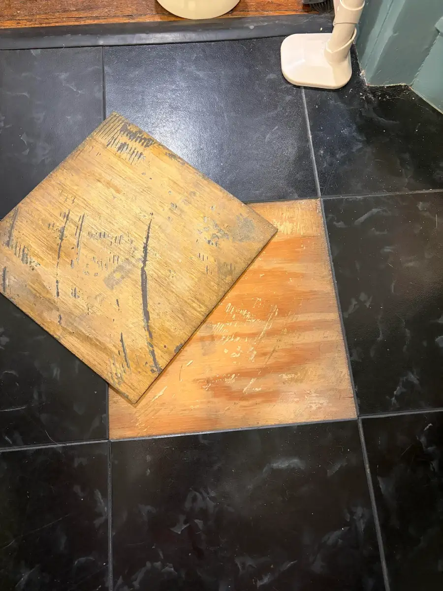 A black tile floor with one loose tile revealing a wooden surface underneath; the removed tile is resting on the floor, showing scratches and wear.