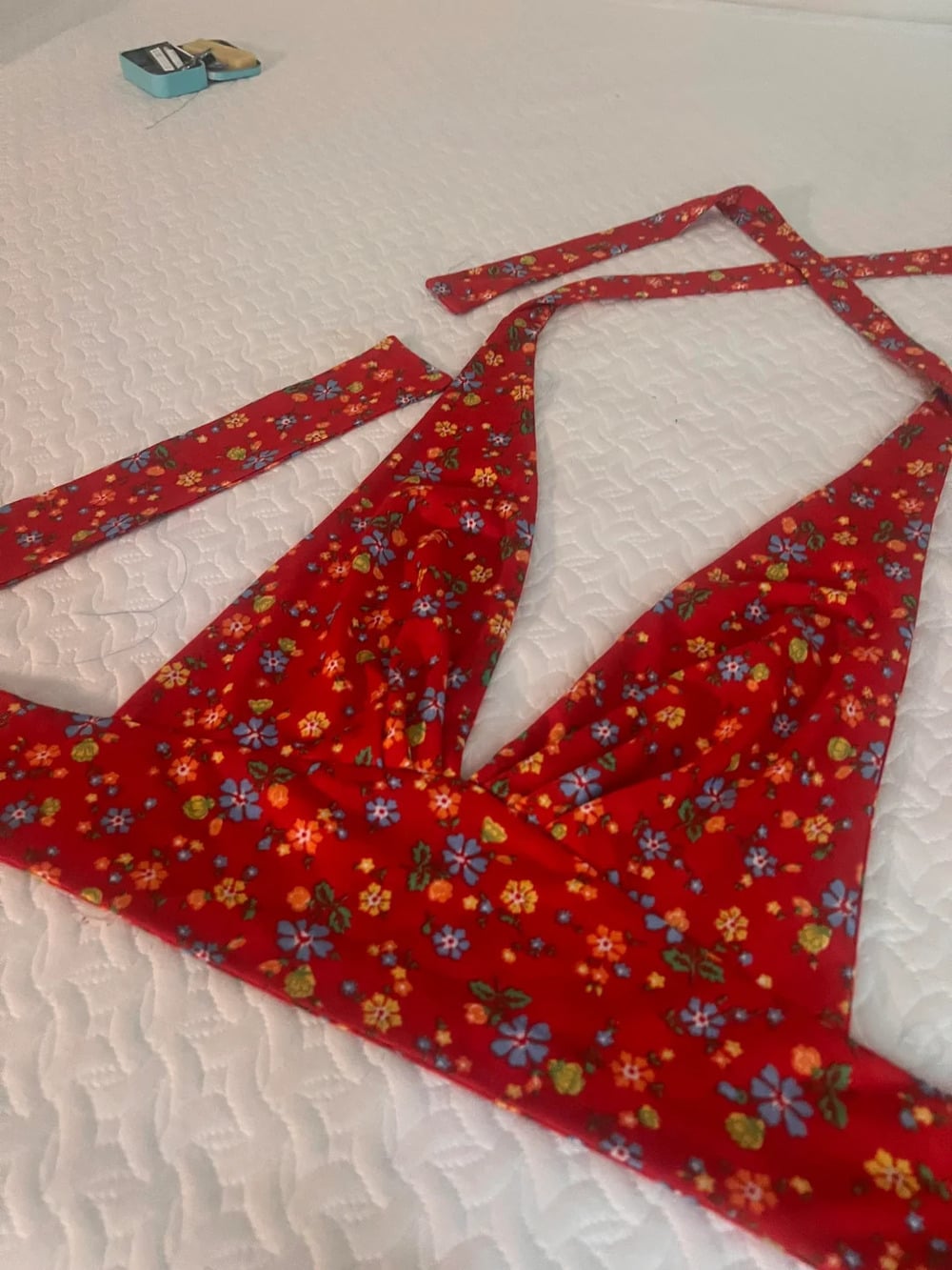 A red floral-patterned bikini top with straps is laid out on a white textured surface. A small blue box is visible in the background.