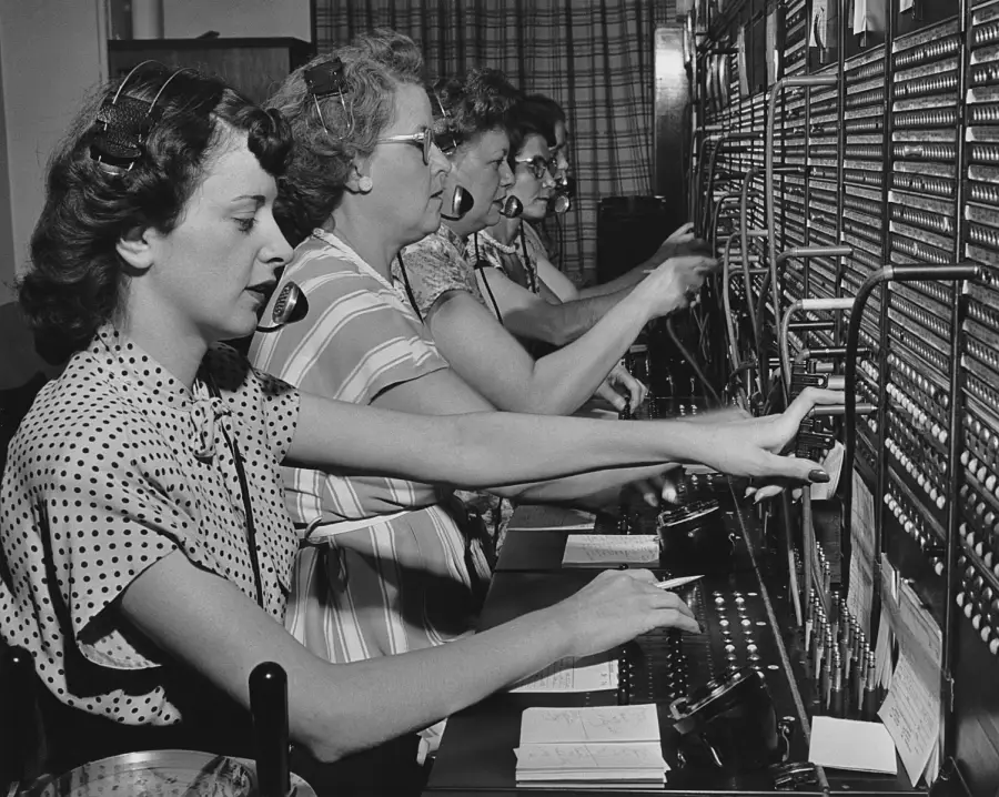Five women wearing headsets and patterned dresses work at a vintage telephone switchboard, connecting calls by plugging and unplugging cables. Curtains cover the window in the background.