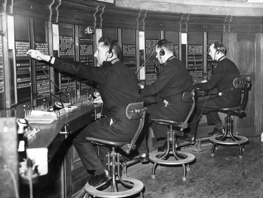 Three men in uniforms sit on tall chairs, operating a vintage telephone switchboard with numerous cables and plugs in an old-fashioned office setting.