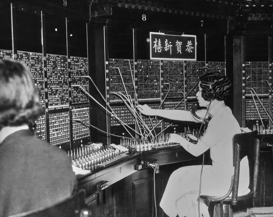 A woman operates a vintage telephone switchboard, connecting cables to different ports. Another person sits nearby. A sign with Chinese characters hangs above the switchboard.