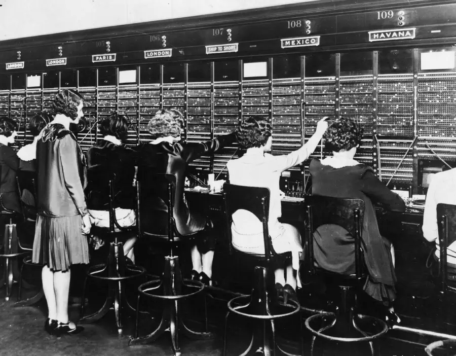 A group of women sit and operate a vintage telephone switchboard, connecting calls to various cities labeled above each station, such as London, Paris, Mexico, and Havana.
