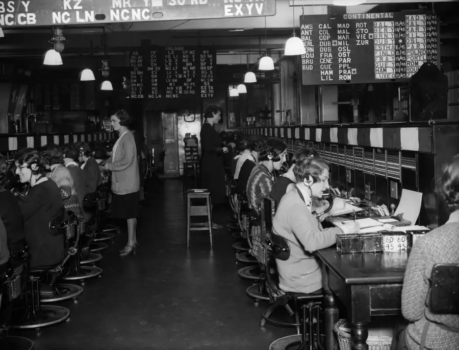 Black-and-white photo of women working at a large telephone switchboard, wearing headsets. Two women stand overseeing the operators. Illuminated boards with destination codes hang overhead.