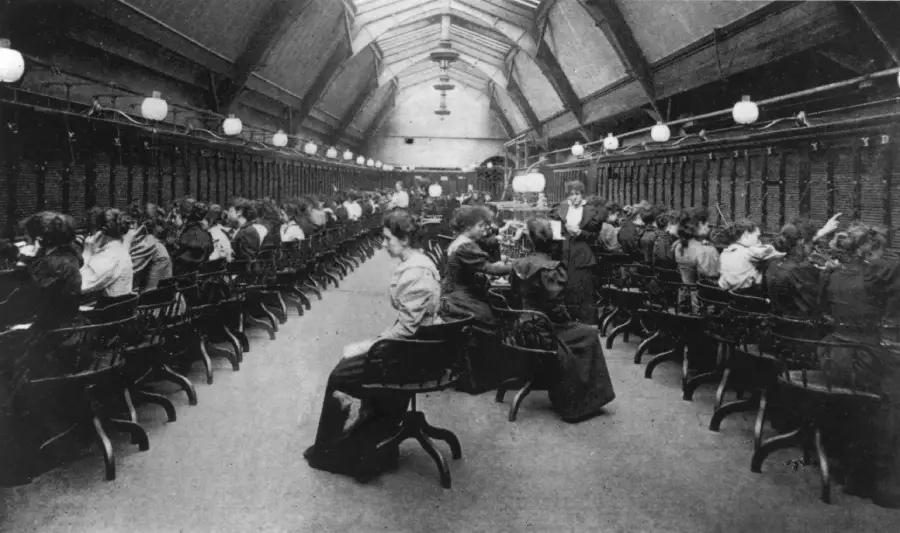 Black and white photo of women sitting in rows at desks, operating telephone switchboards, inside a large, high-ceilinged room with hanging lights.