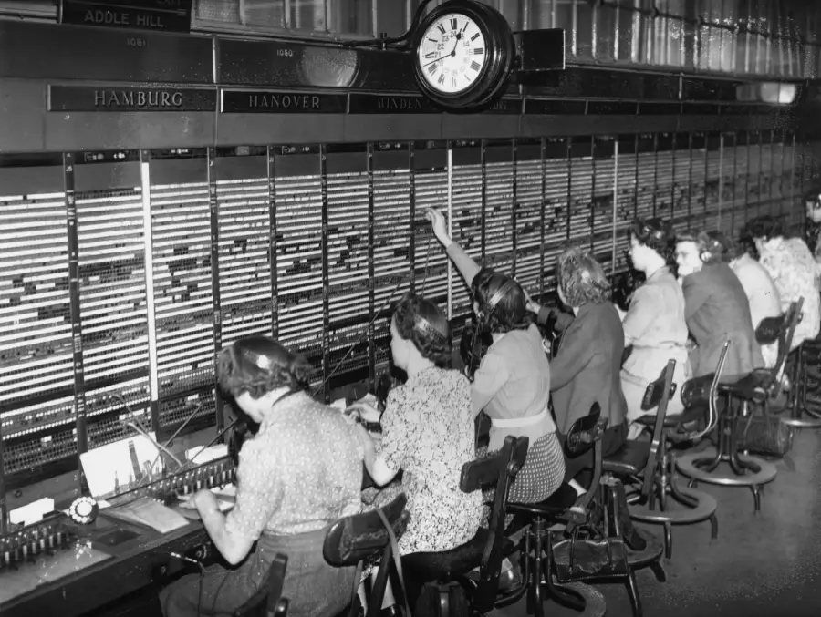 A group of women wearing headsets work at a large telephone switchboard, connecting calls by plugging wires. A clock and city names are visible above the switchboard.