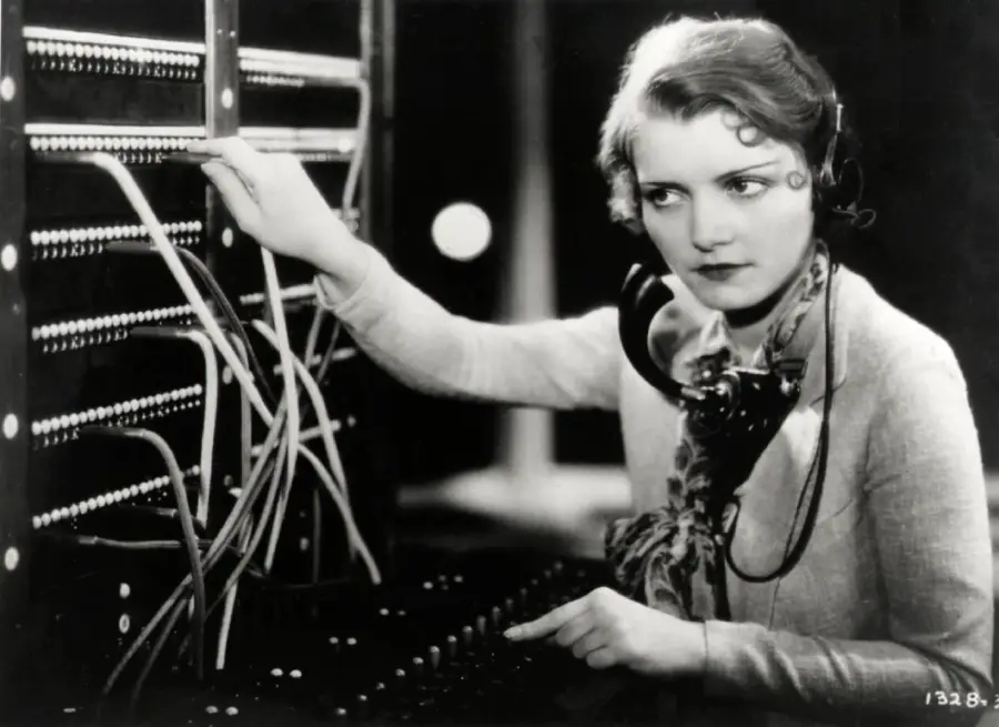 A woman in vintage clothing operates a telephone switchboard, wearing a headset and connecting cables, in a black-and-white photo.