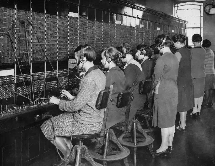 A group of women in vintage clothing work at a large telephone switchboard, connecting calls with cables, wearing headsets in a busy office.