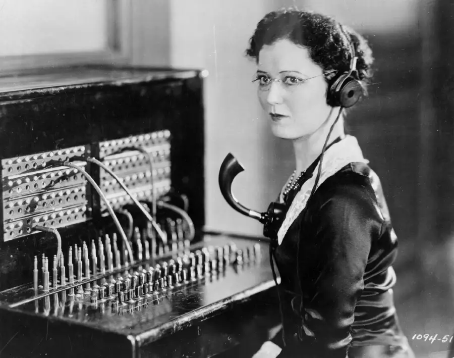 A woman in vintage clothing sits at a telephone switchboard, wearing a headset and glasses, holding a telephone receiver as she connects calls using cables. The image is black and white, evoking an early 20th-century office setting.