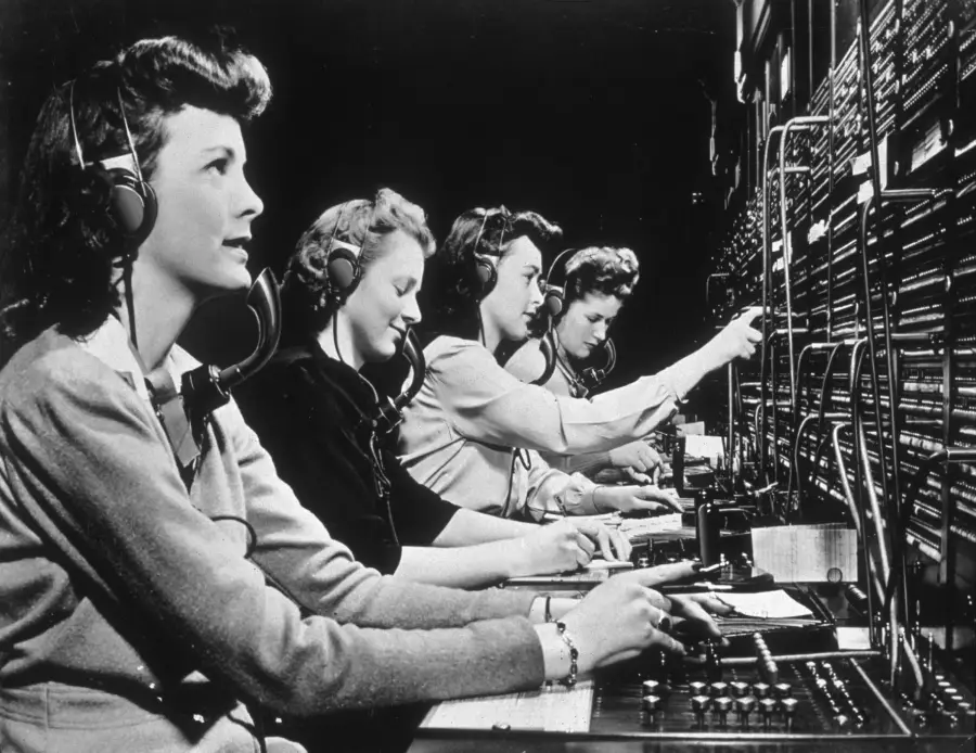 Four women wearing headsets operate a large old-fashioned telephone switchboard, plugging and unplugging cables as they work in a row. The image is in black and white.