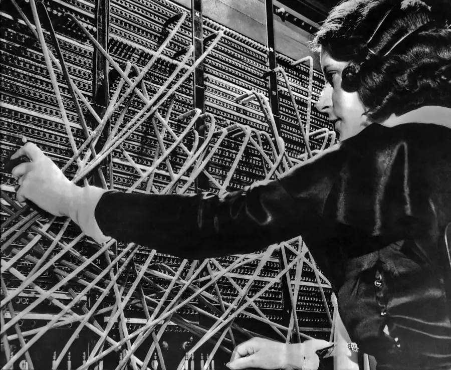 A woman operates a vintage telephone switchboard, connecting calls by plugging in multiple cables.