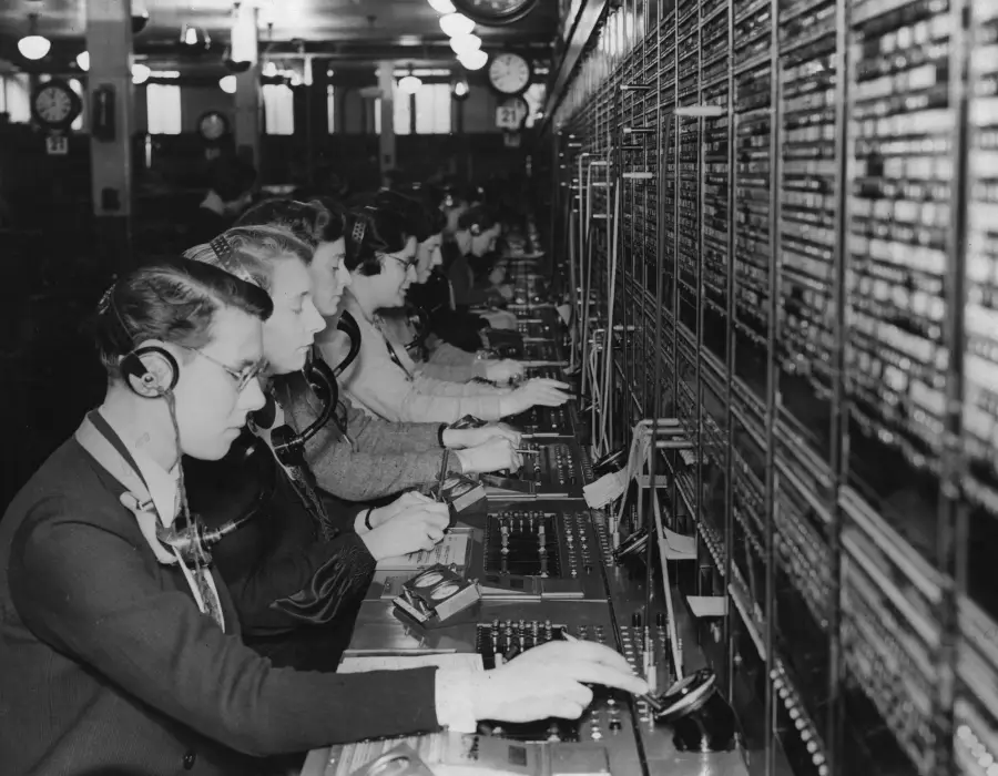 A row of telephone operators wearing headsets work at a large switchboard, connecting calls in a busy office filled with equipment and clocks on the wall.