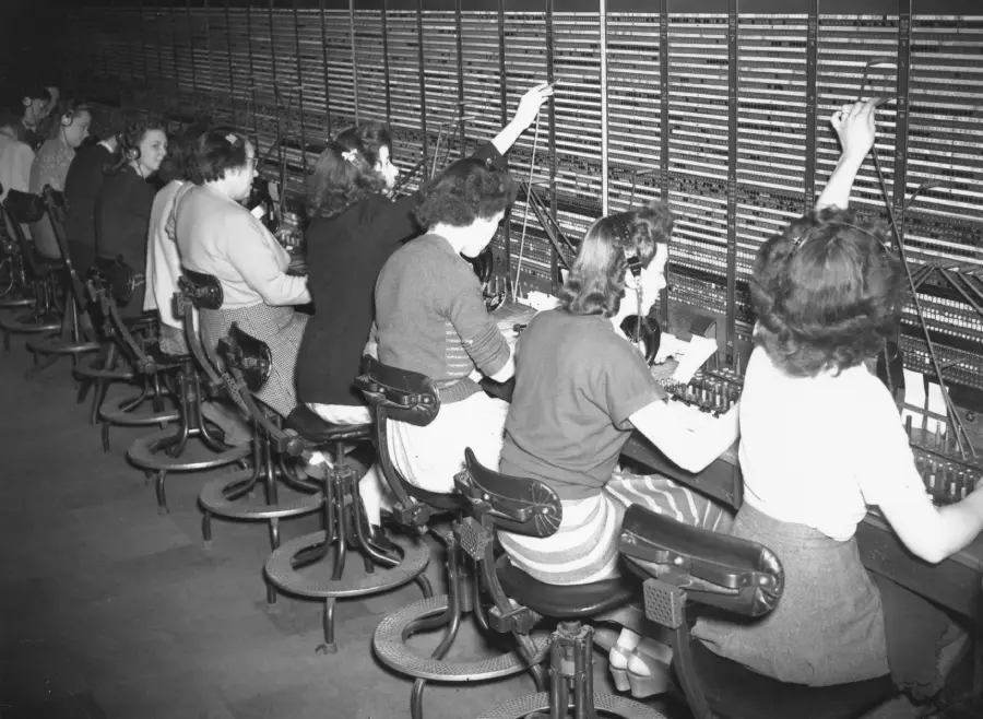 Black-and-white photo of women sitting in a row at a telephone switchboard, wearing headsets and connecting calls with wires.