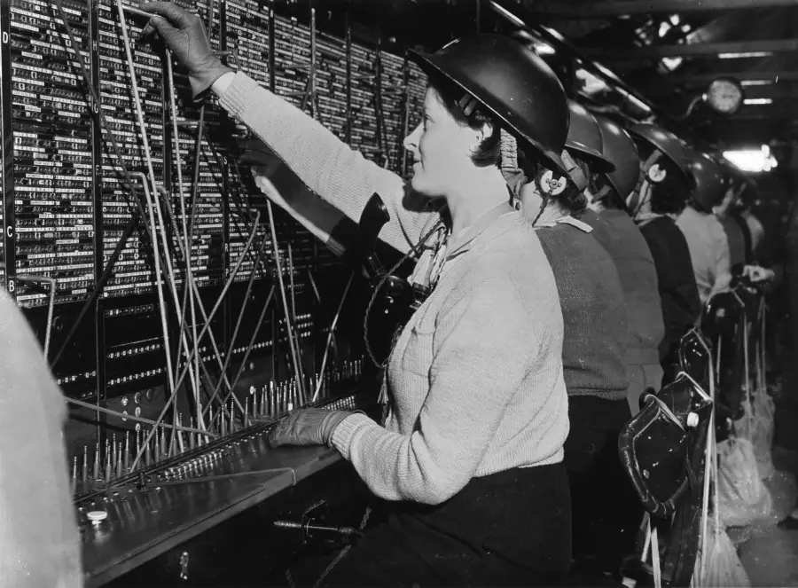 Women in hard hats work at a large, busy telephone switchboard, connecting calls with cords. They sit in a row, focused on their tasks, wearing sweaters and headsets in a black-and-white industrial setting.