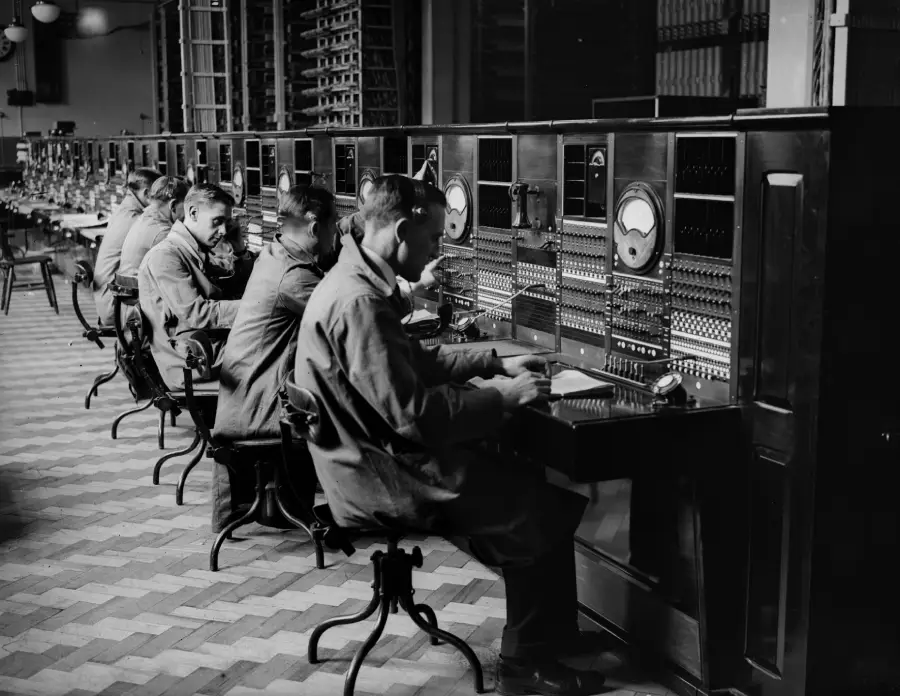 Several operators sit in a row at large, vintage telephone switchboards, wearing uniforms and working with cables and dials in a black-and-white, early 20th-century setting.