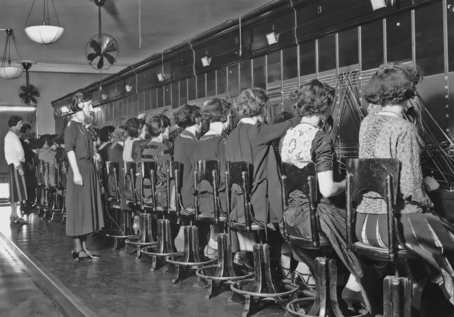 A row of women sits at a long switchboard operating telephones, while two women stand nearby in a vintage black-and-white setting, likely from the early 20th century.