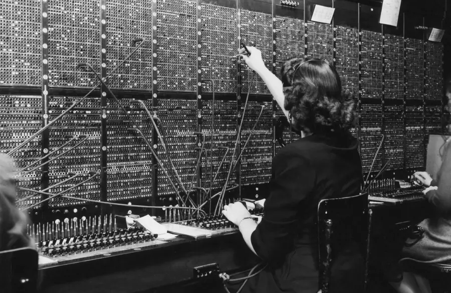 A black-and-white photo of two women operating a vintage telephone switchboard, connecting calls using cables and plugs.