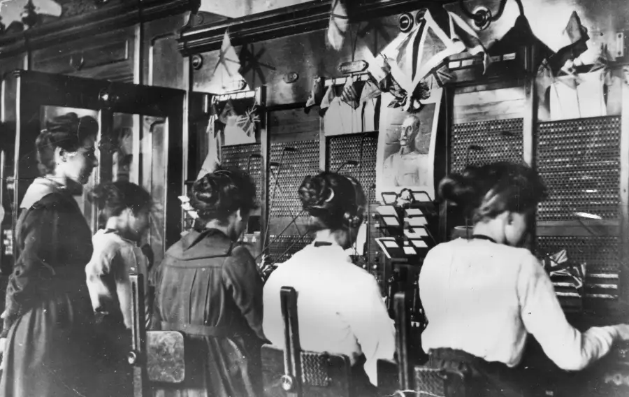 Four women sit and one stands at an old-fashioned telephone switchboard, connecting calls with wires. The workspace has papers, photos, and decorations pinned above the switchboard. The image is black and white.