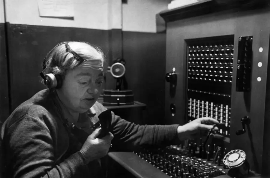 An older woman operates a vintage telephone switchboard, wearing a headset and holding a microphone while adjusting switches on the panel.
