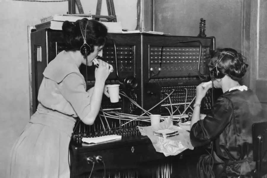 Two women sit at a vintage telephone switchboard, wearing headsets and connecting calls with cords. They appear to be on a break, eating and drinking while still at their workstations.