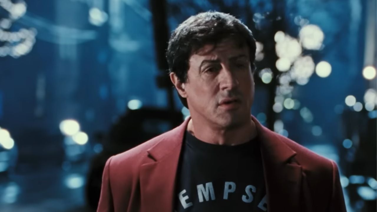 A man with short dark hair wears a red blazer over a dark shirt, standing outside at night with blurred streetlights and trees in the background.
