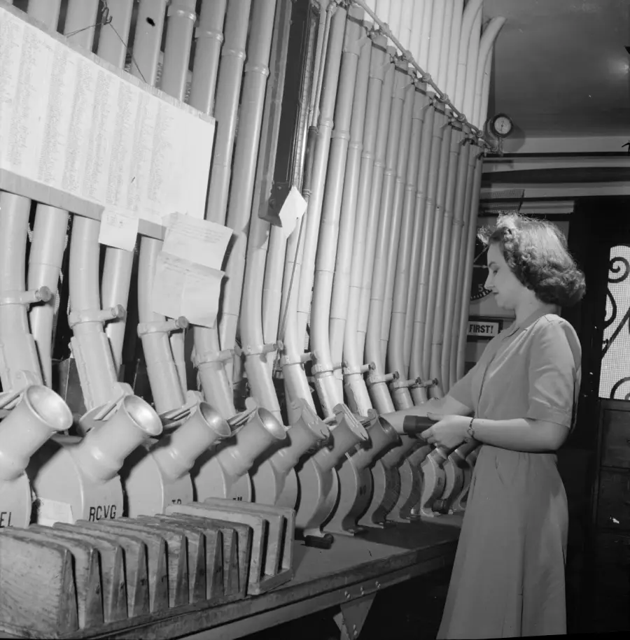 A woman stands at a row of large, labeled pneumatic tubes in an office, inserting or retrieving documents. Papers and lists are attached above the tubes. The scene is lit brightly, suggesting daytime.